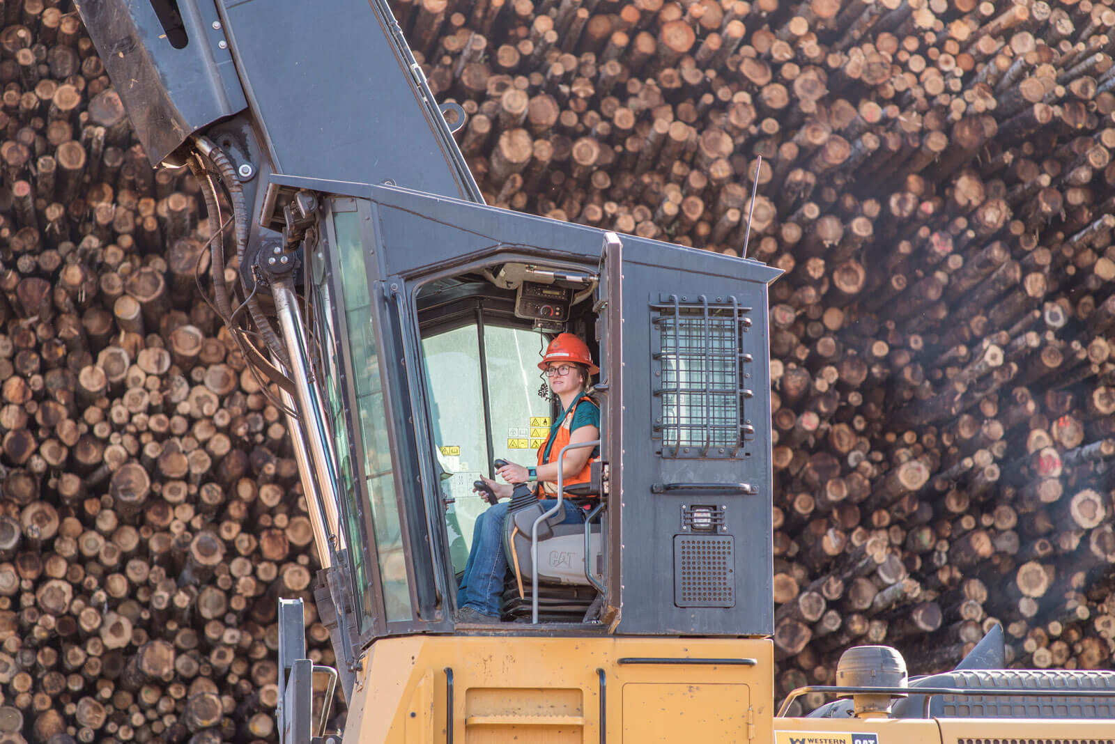 A woman operating large machinery in front of a large pile of logs.